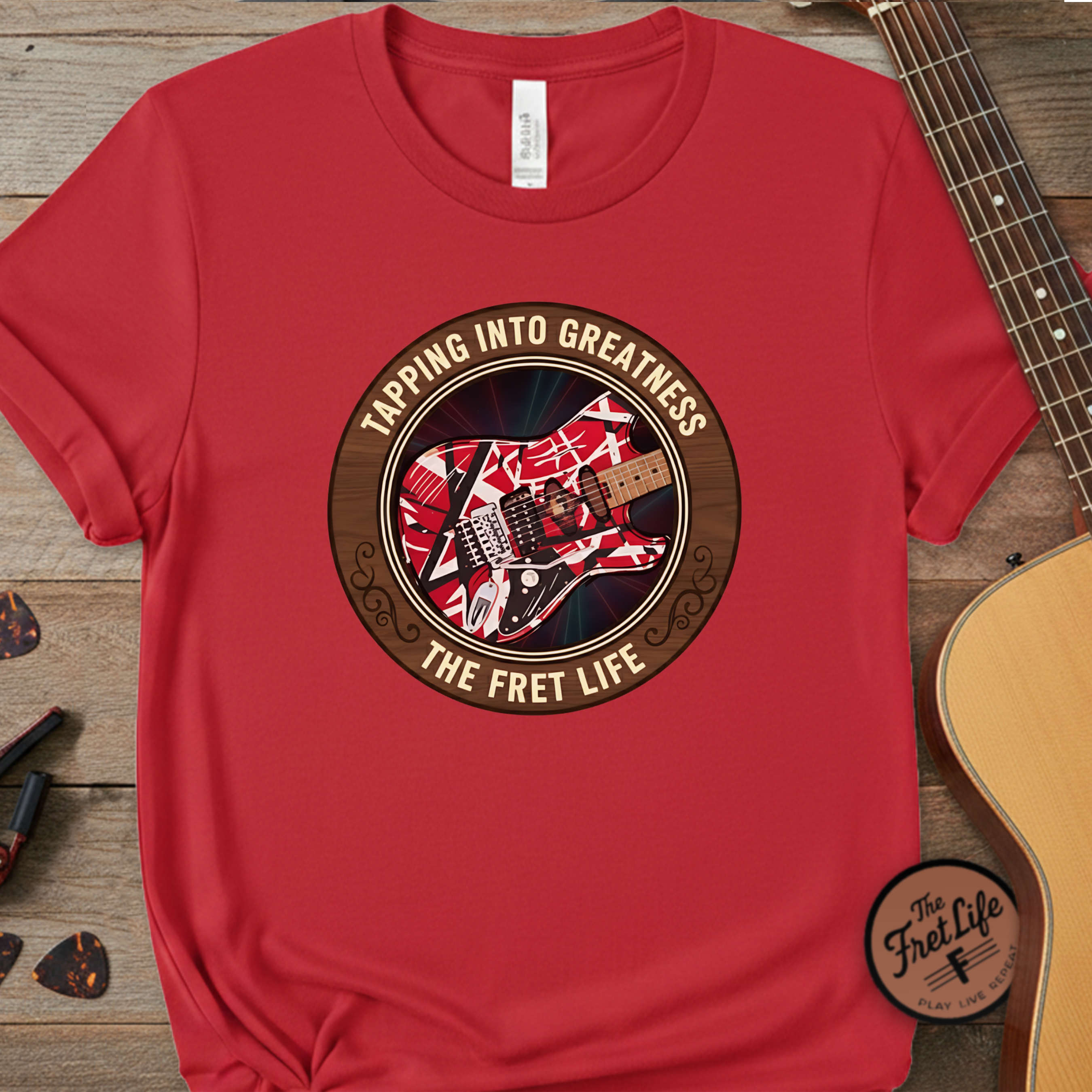 Red t-shirt with guitar graphic and text 'Tapping into Greatness, The Fret Life' on a wooden surface with a guitar.