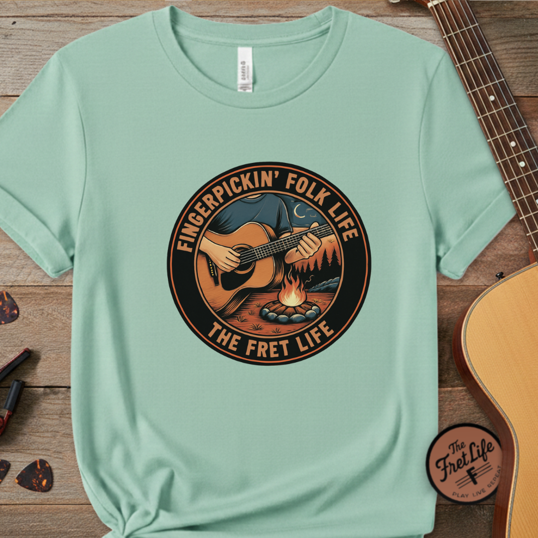 Light green t-shirt with 'Fingerpickin' Folk Life The Fret Life' graphic on a wooden surface with a guitar.