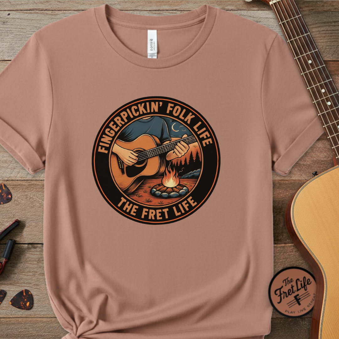 Brown t-shirt with 'Fingerpickin' Folk Life' graphic on a wooden surface with a guitar.