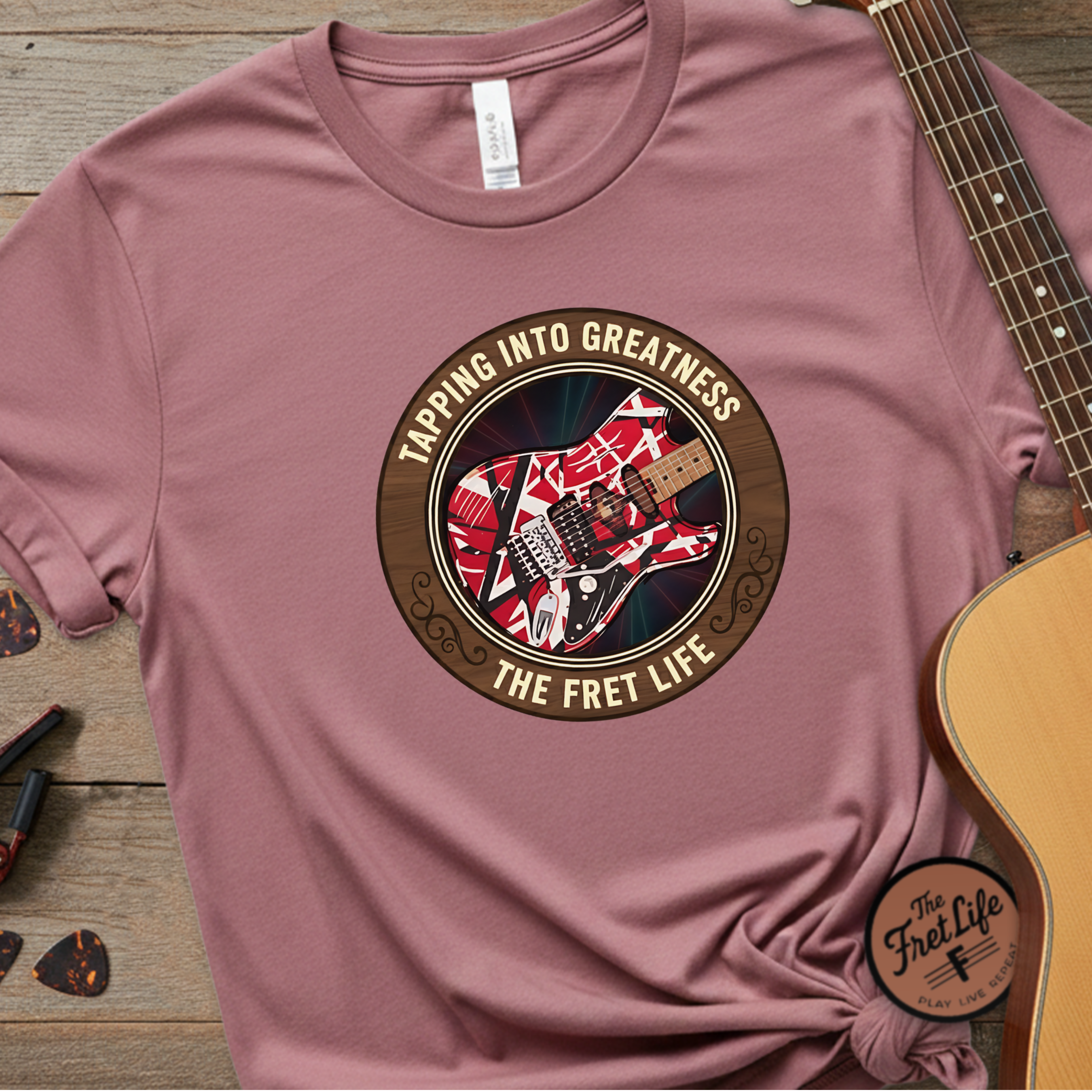 Pink t-shirt with guitar graphic and text 'Tapping Into Greatness, The Fret Life' on a wooden surface with a guitar.