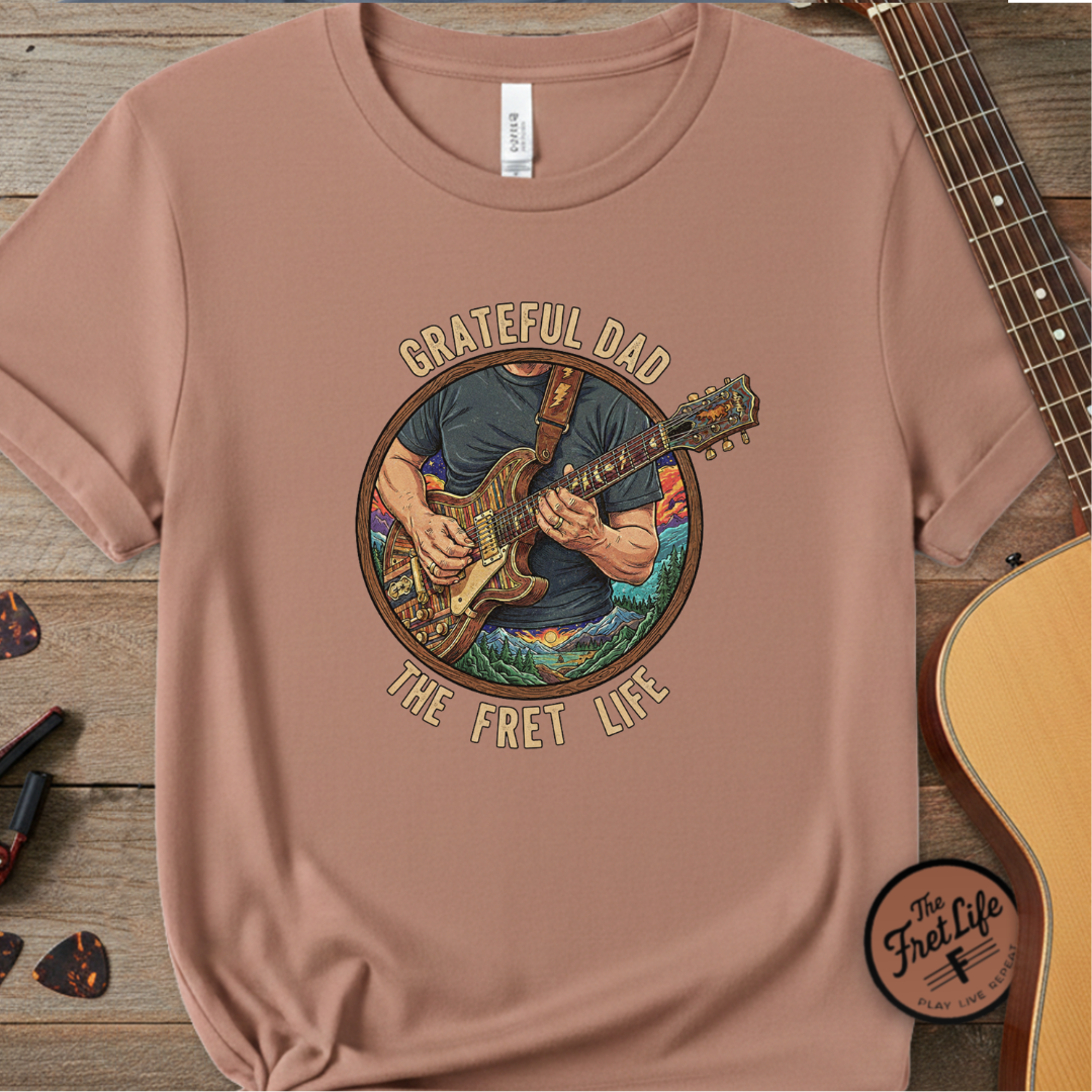 Brown t-shirt with 'Grateful Dad The Fret Life' graphic on a wooden surface with a guitar.