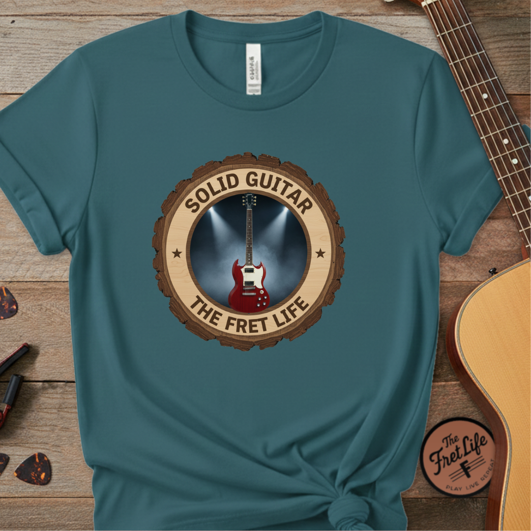 Teal t-shirt with 'Solid Guitar The Fret Life' design on a wooden surface with a guitar.