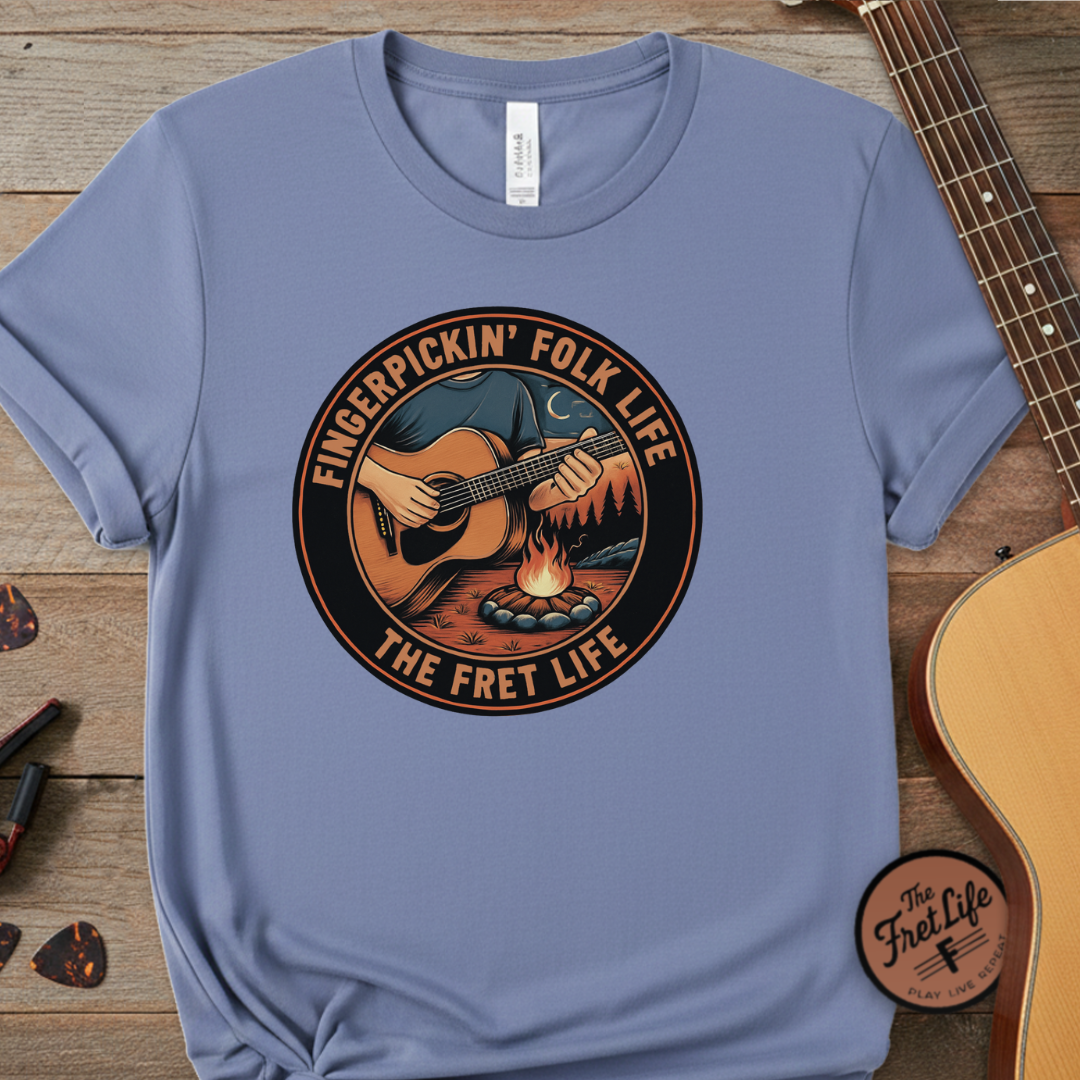 Blue t-shirt with 'Fingerpickin' Folk Life' graphic on a wooden surface with a guitar.