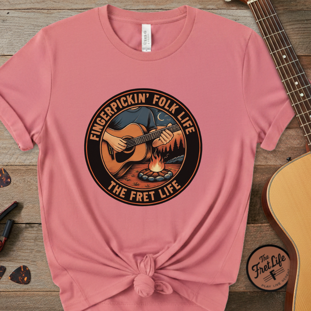 Pink t-shirt with 'Fingerpickin' Folk Life The Fret Life' design on a wooden surface with a guitar.