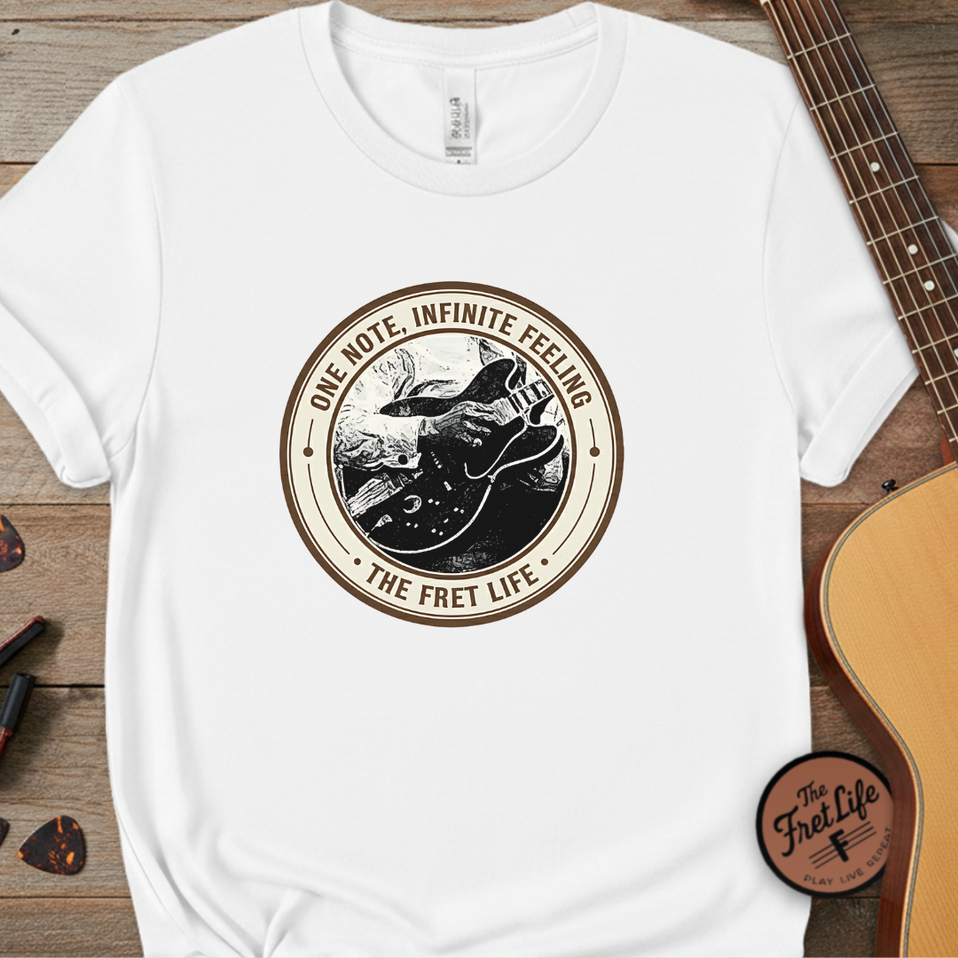 White t-shirt with 'The Fret Life' graphic design on a wooden surface with a guitar.