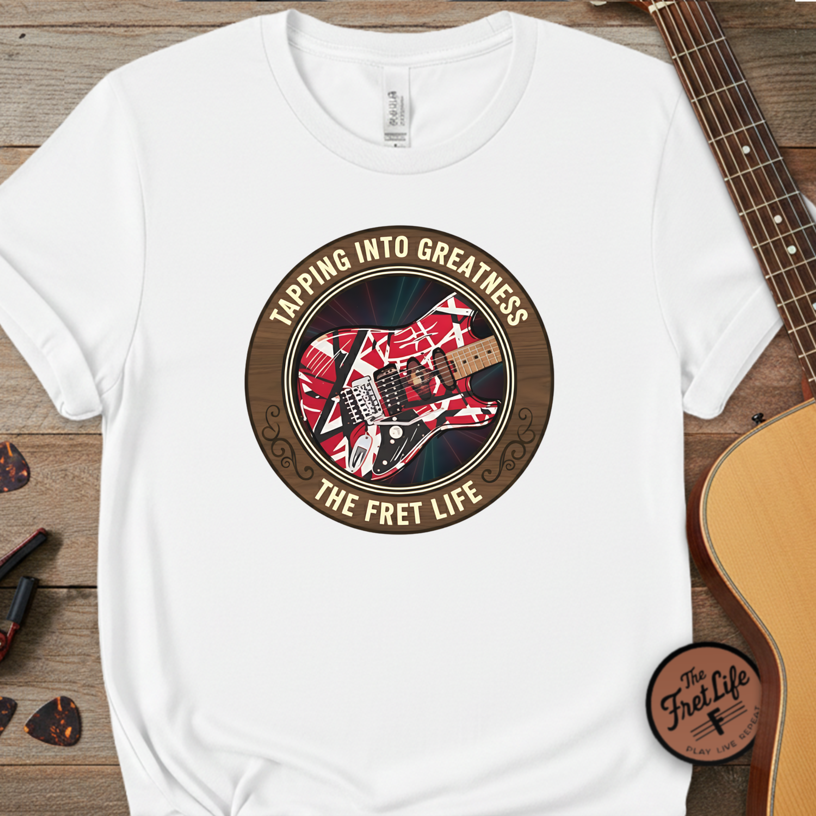 White t-shirt with guitar graphic and text 'Tapping Into Greatness, The Fret Life' on a wooden surface with a guitar.