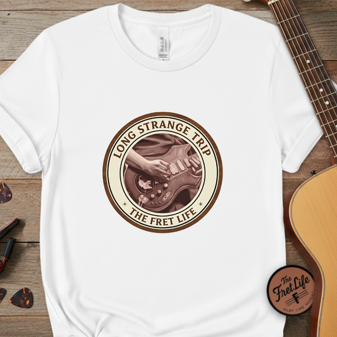 White t-shirt with 'Long Strange Trip: The Fret Life' graphic on a wooden surface with a guitar.