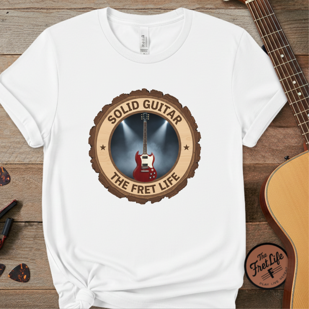 White t-shirt with 'Solid Guitar The Fret Life' design on a wooden surface with a guitar.