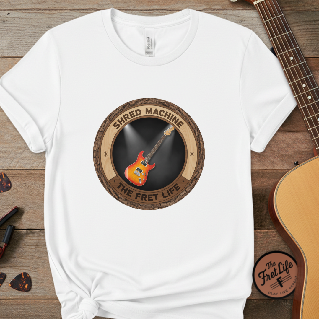 White t-shirt with guitar graphic and 'Shred Machine' text on a wooden surface with guitars.