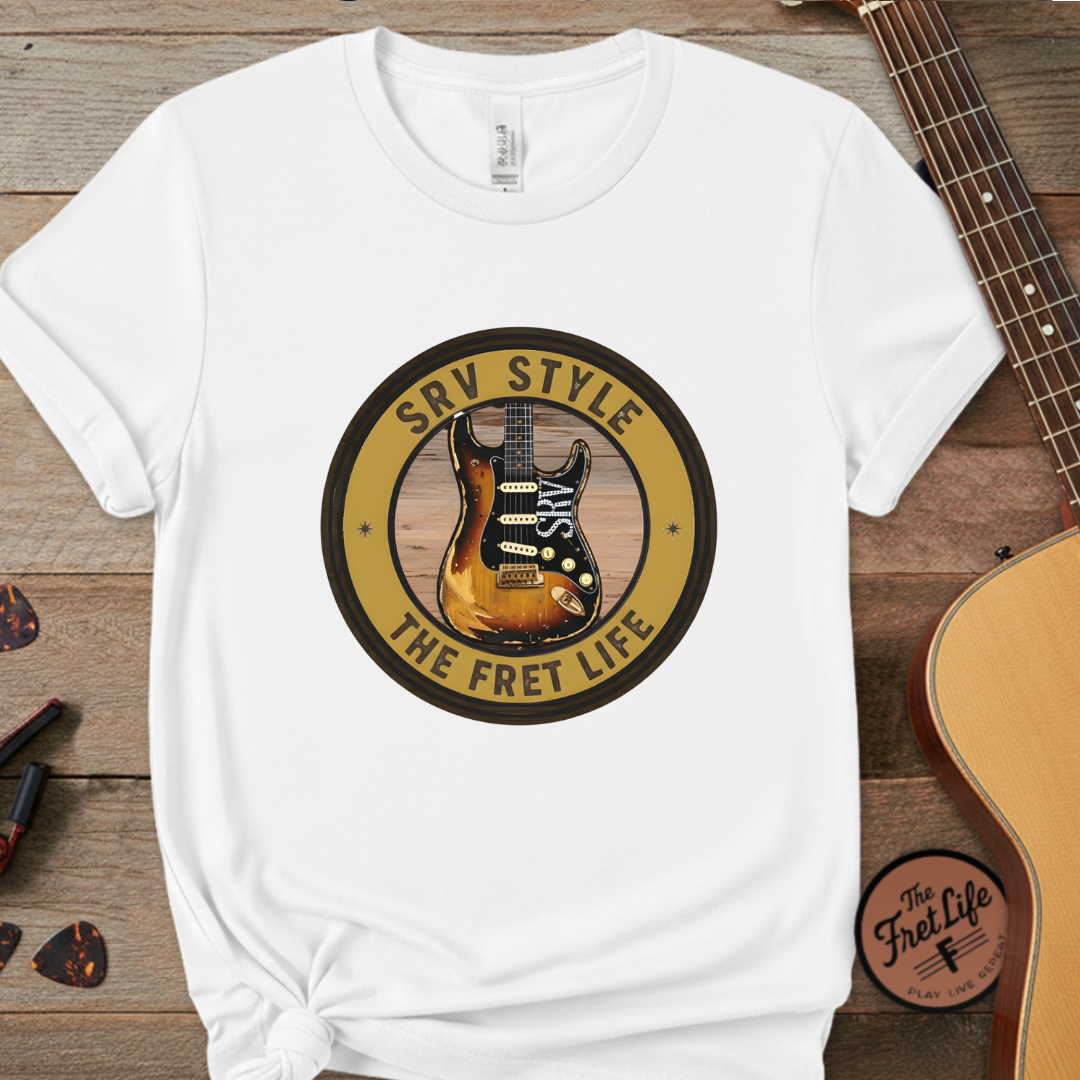 White t-shirt with guitar graphic and text on a wooden surface with guitars and picks.