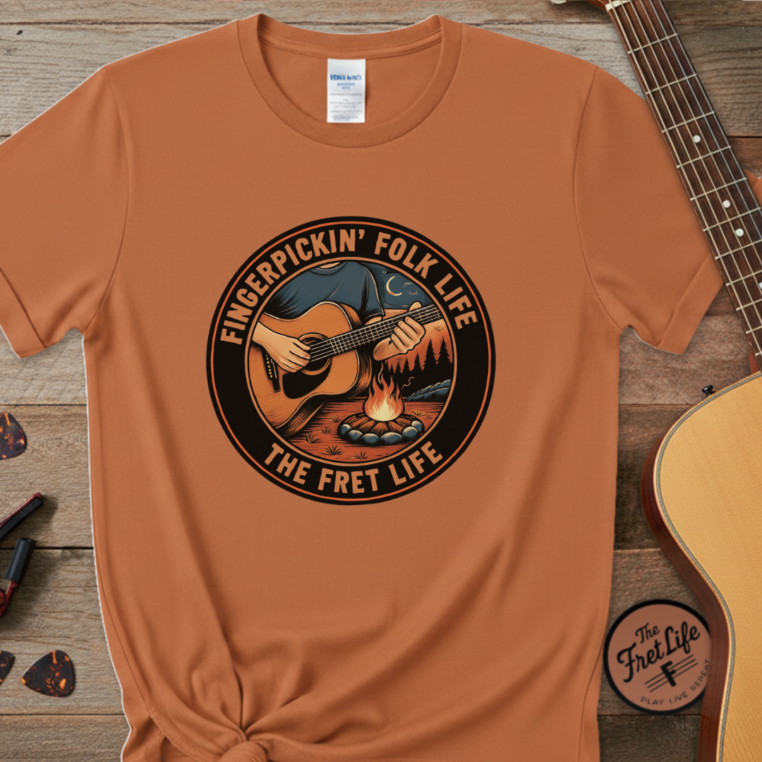 Brown t-shirt with 'Fingerpickin' Folk Life' graphic on a wooden surface with a guitar.