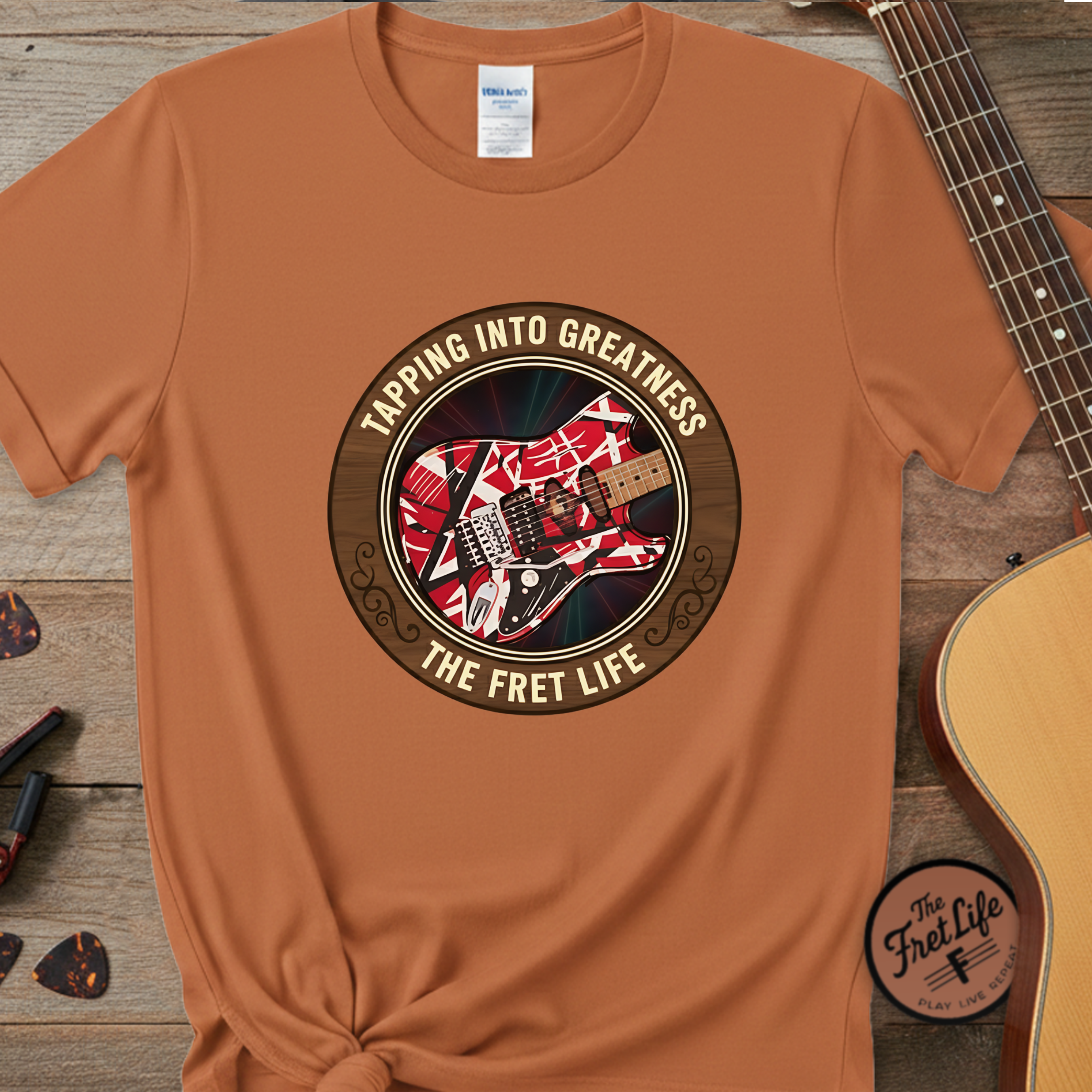 Brown t-shirt with 'The Fret Life' graphic design on a wooden surface with a guitar.