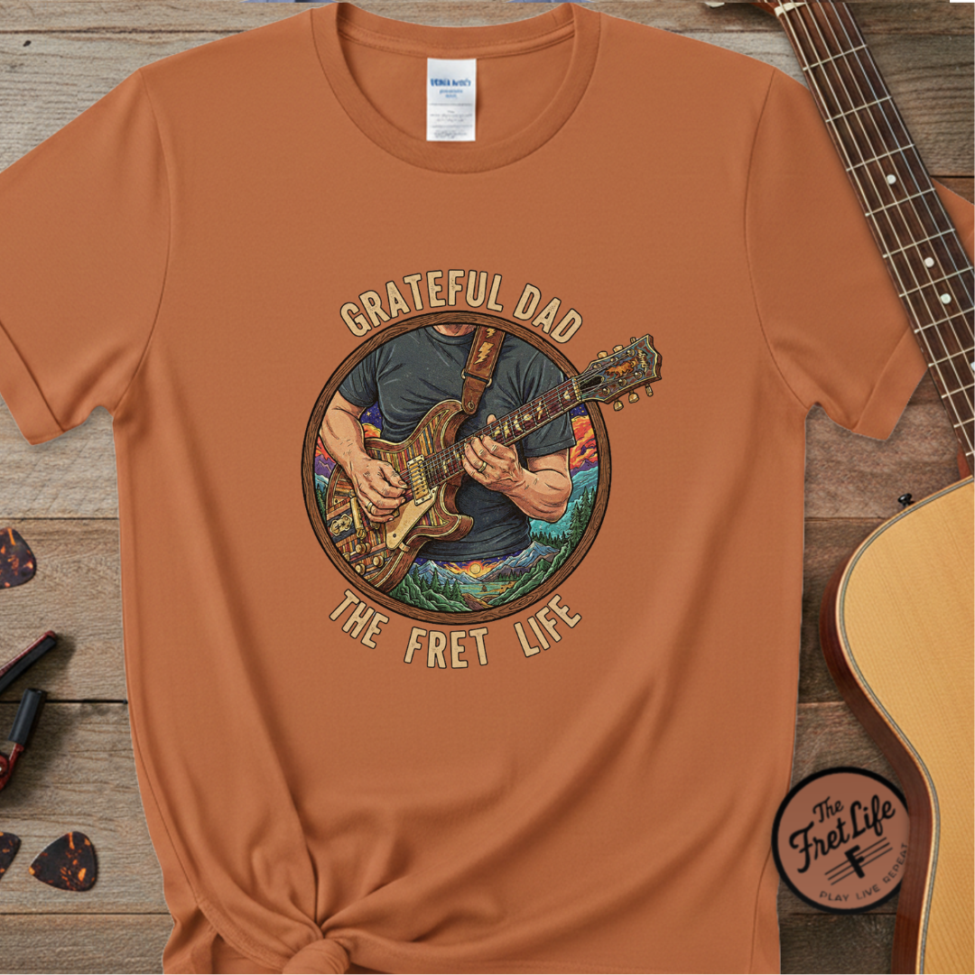 Brown t-shirt with 'Grateful Dad The Fret Life' graphic on a wooden surface with a guitar.